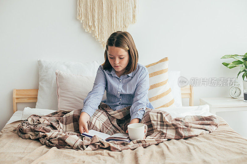 女孩拿着一杯茶，读着一本书，休息时坐在床上。舒适的居家季节，意境概念