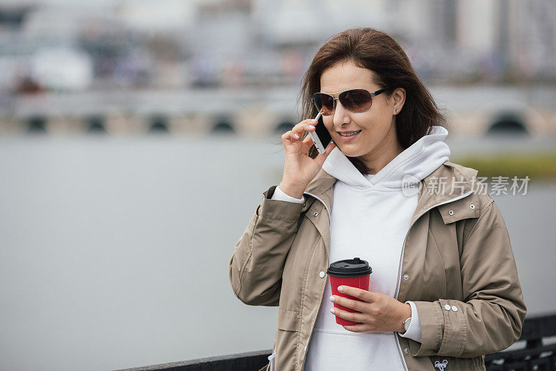 美丽的白种女人戴着墨镜走在路堤上，手里拿着咖啡，用手机说话，选择性地专注。