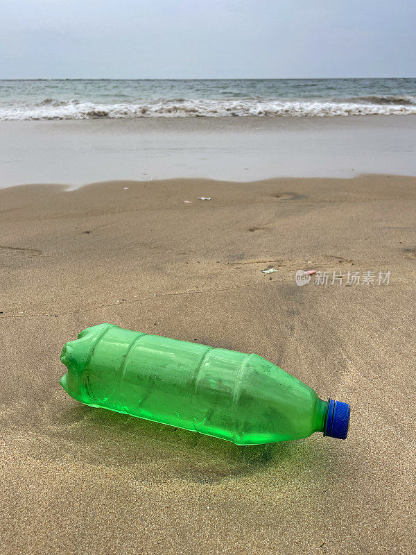 空的，蓝色盖子的绿色塑料饮料瓶被冲到退潮水边缘的沙滩上，破碎的海浪冲刷着海洋垃圾和岸上的污染，乱扔的沙子，肮脏的海滩，关注前景，复制空间