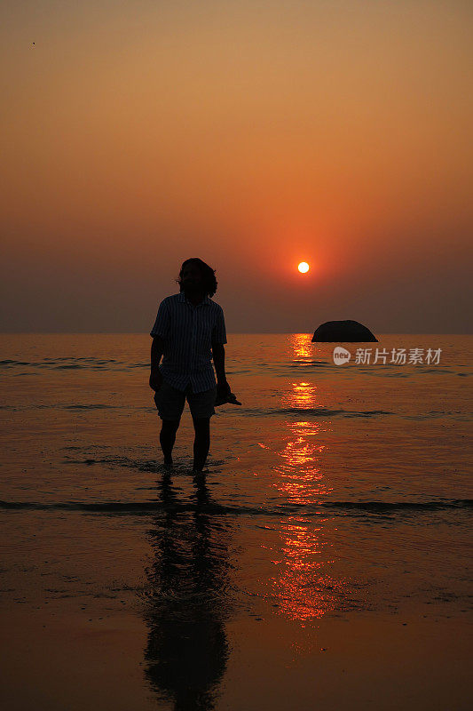 印度果阿的Palolem，印度果阿，度假游客赤脚走在海滩上，手里拿着鞋子在水里溅起水来，橙色的日落，看着相机，聚焦在前景