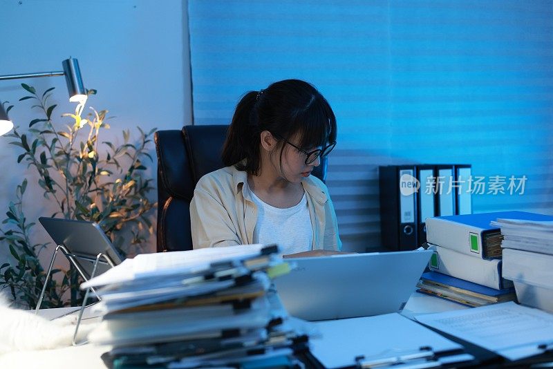 年轻的亚洲女商人在深夜工作。晚上，她坐在漆黑的家庭办公室里，忙着检查文件，不得不在截止日期前把工作送出去。