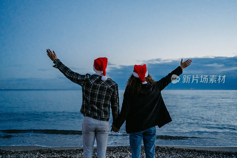 希腊，一对戴着圣诞帽的年轻夫妇在黎明时分欣赏着海岸线的景色