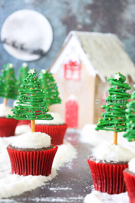 雪景圣诞图片，批量自制，圣诞树设计巧克力纸杯蛋糕排列路径通过糖粉雪姜饼屋，圣诞老人雪橇和驯鹿剪影飞过月亮的背景，重点在前景