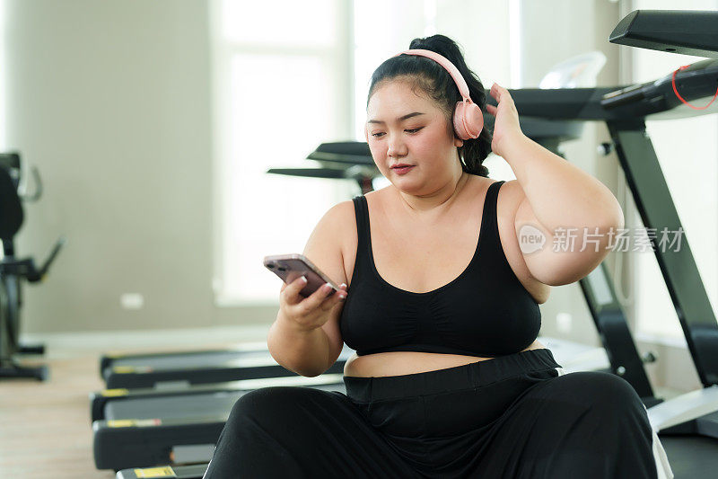 胖乎乎的亚洲女性在健身房锻炼，穿着运动服的女性在休息时愉快地使用智能手机，体现了休闲与健身的平衡。舒舒服服地坐在跑步机上，快乐就会到来