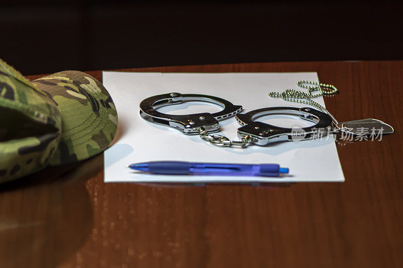 木桌上放着一顶迷彩军帽、一对军章、金属手铐和一支写着一张纸的蓝色钢笔。理念:执法，军事权威。