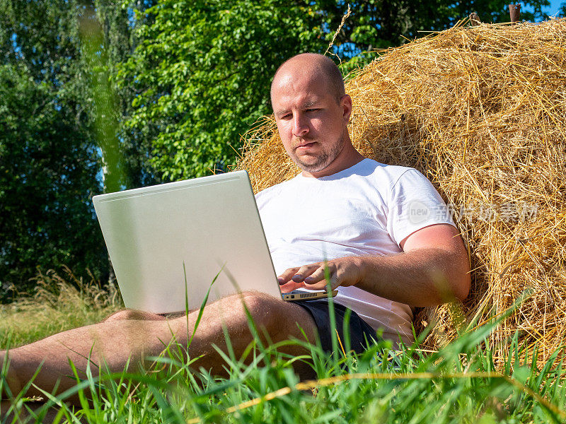 一位身穿t恤和短裤的中年男子正躺在黄色的干草堆上，手里拿着一台灰色的笔记本电脑。远程工作，培训。农村地区。他脸上若有所思的表情