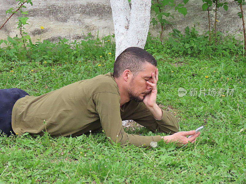 英俊的胡子男子躺在树下使用他的智能手机