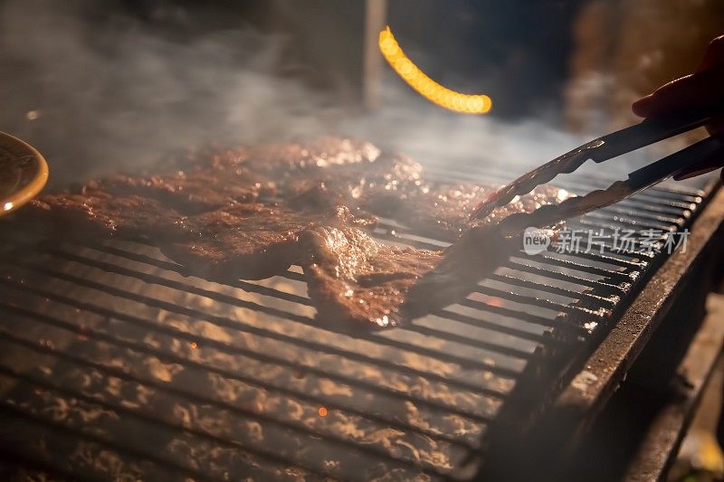 晚上，一个烧烤架，美味多汁的牛排在篝火上烤着，一只手拿着钳子，把肉翻过来。特写,柔软的焦点。烟是强调