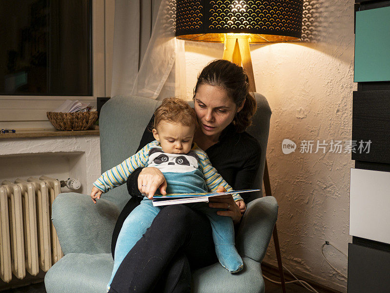 年轻的母亲在舒适的椅子上为小儿子读睡前故事