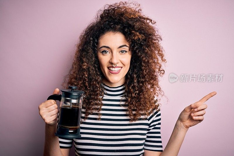 年轻美丽的女人与卷发和穿洞做咖啡拿着法国咖啡机非常高兴的指向与手和手指的一边