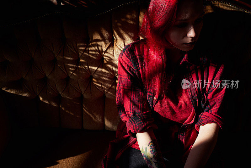 年轻的红头发女人在红色的衬衫上纹身和化妆坐在沙发上在昏暗的沙龙和看着别处沉思