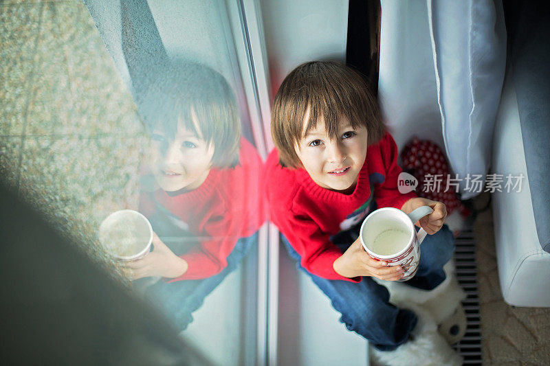 可爱的学龄前儿童，小男孩，喝着牛奶，坐在窗户上