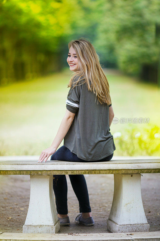 一个厚脸皮的十几岁的女孩坐在石凳上大笑，看着她的肩膀与模糊的绿色背景