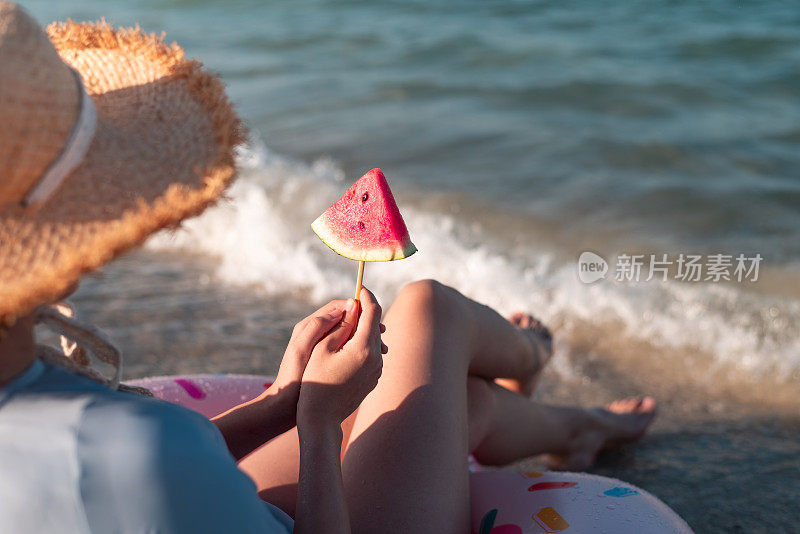 一个女人坐在沙滩上吃着西瓜，同时坐在一个漂浮的甜甜圈上