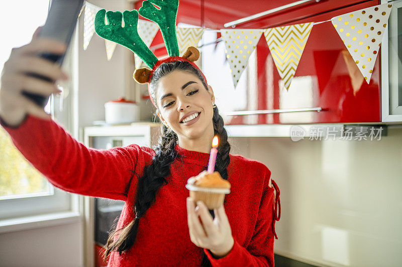 戴着圣诞老人帽的年轻女士正在许愿，拿着带蜡烛的纸杯蛋糕，在厨房里视频通话