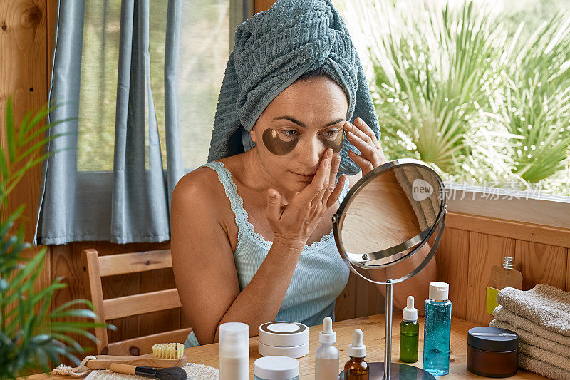 护肤习惯。中年妇女抚摸她的脸上有皱纹和水凝胶下眼罩和看着镜子。提升眼下抗皱面膜。胶原蛋白面膜，护肤和水疗概念。