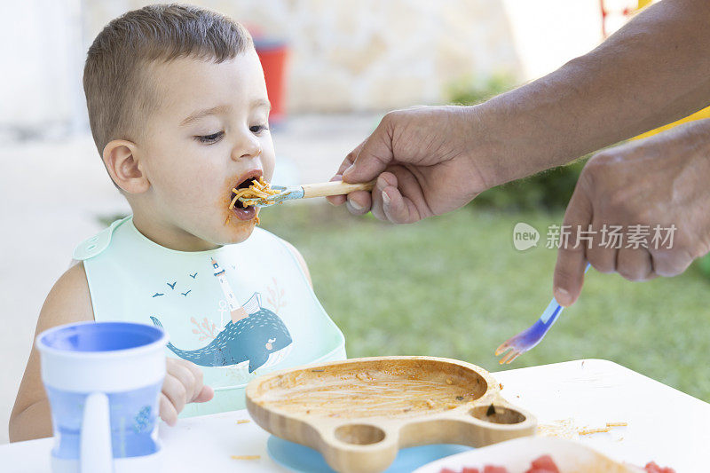 一个可爱的小男孩在他爷爷的帮助下吃饭。一个小男孩坐在花园里的椅子上吃东西。