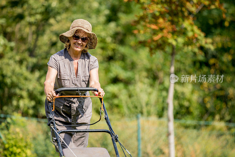 成熟的女人在花园里工作，用割草机割草