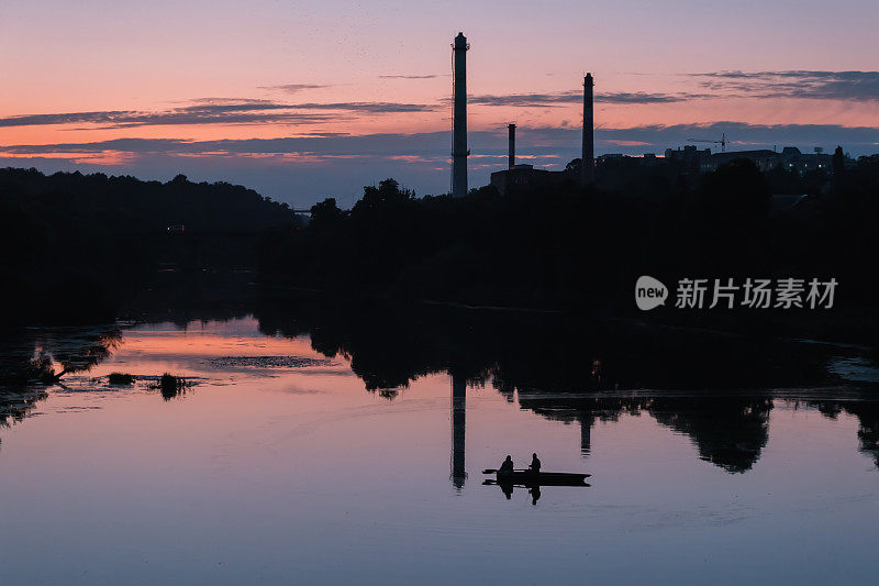 夕阳下的河上，两名男子在船上，背景是工厂的管道
