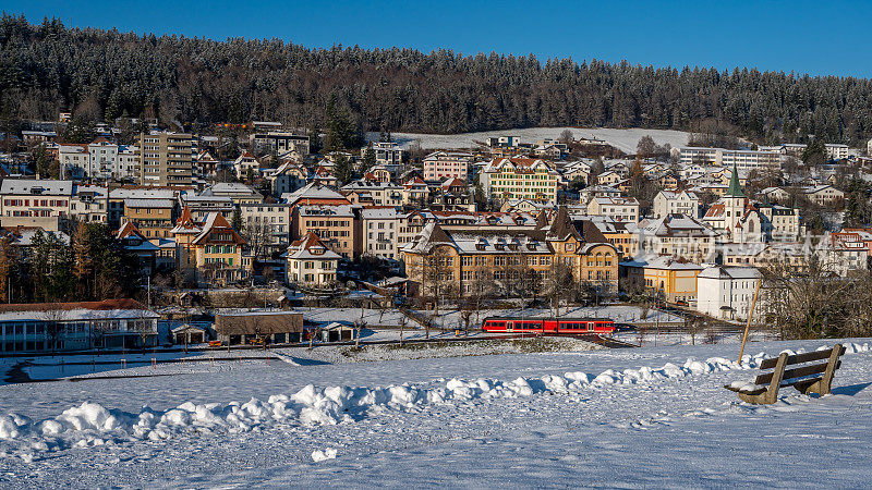 有雪的冬季景观。阳光灿烂的日子里，红色火车穿过Tramelan村。