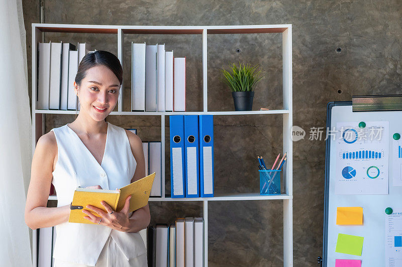 亚洲女商人举着一块有图表的板子展示商业数据，图表手拿着一个黄色的笔记本，而商业文件则放在办公室的桌子上，展示商业概念。