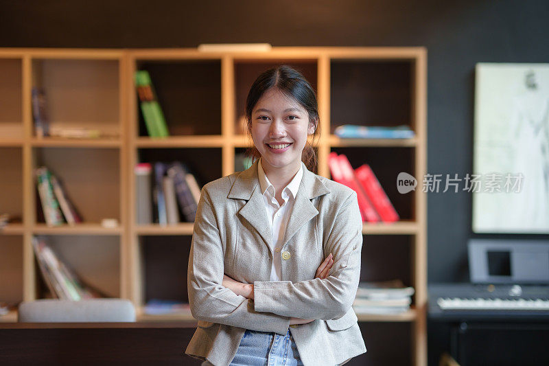 一个迷人的，美丽的亚洲女商人站在她的办公室里，双臂交叉，微笑着，开心地看着镜头。