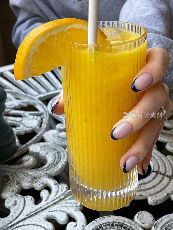 特写照片是一个不知名的女人拿着一杯橙汁和橙片，还有纸吸管，餐厅的露天铁制桌子，重点放在前景上
