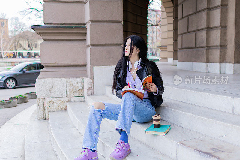 年轻漂亮的女大学生背着双肩包坐在大学楼梯上。她戴着耳机，穿着皮夹克，等待着大学课程的开始。