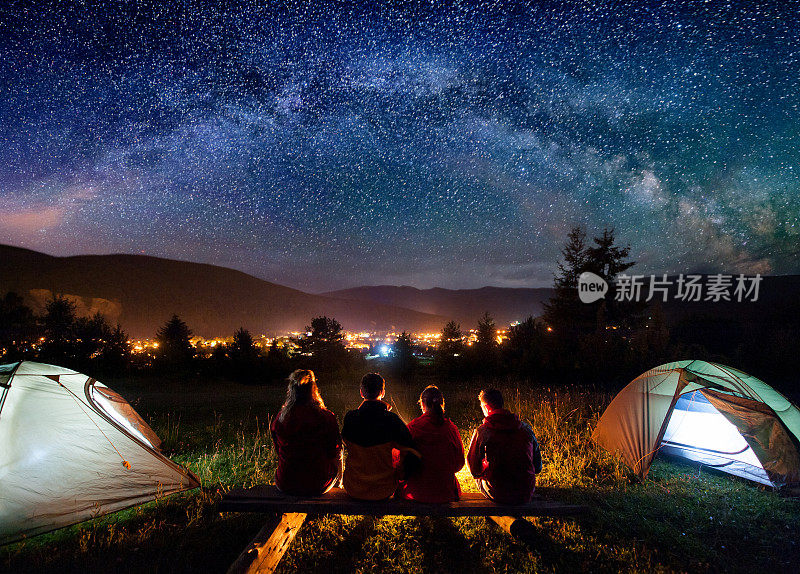 4个人的剪影坐在一个由原木制成的长凳上，一起看火在营地和帐篷旁边的夜晚。背景是星空、银河、群山和发光的小镇。后视