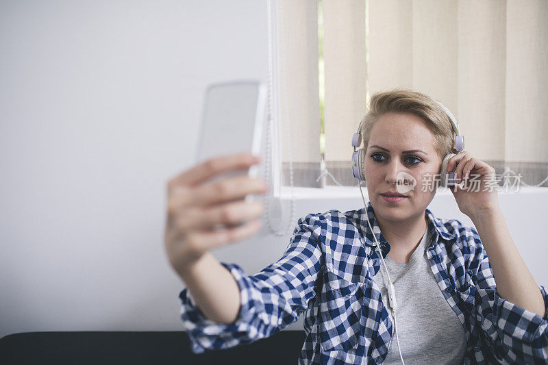 带着耳机的微笑女人在家里用智能手机自拍。
