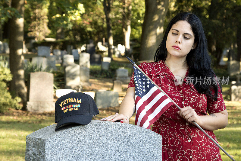 一名年轻女子坐在墓地的墓碑上，凝视着越战老兵的军帽，背景是墓碑