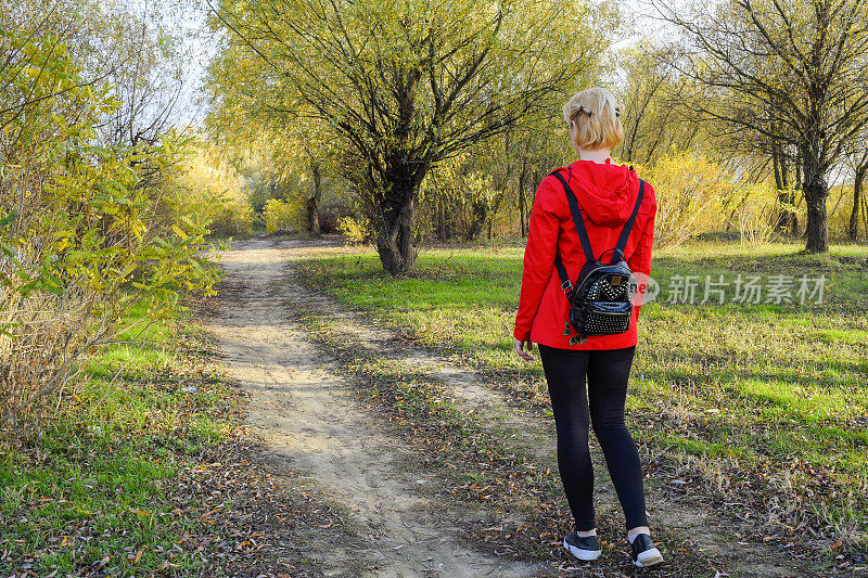 女孩穿着红色的夹克和一个背包沿着路去森林。路的开始。