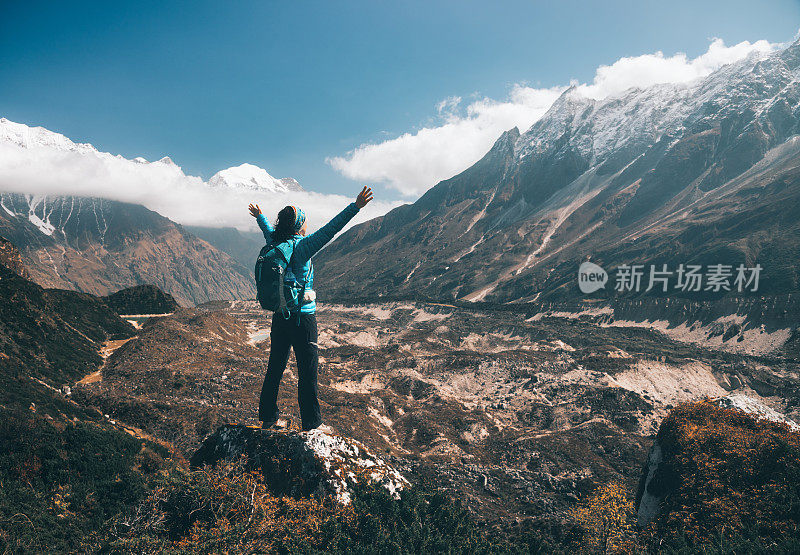 站在山上的年轻女子背着背包，举起双臂，望着群山。风景与快乐的女孩，高山，蓝天与云朵在秋天的尼泊尔。旅行。徒步旅行在喜马拉雅山脉