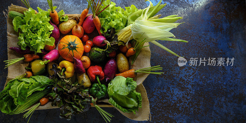 许多彩色的对比色沙拉蔬菜在麻袋材料坐在一个木制蔬菜托盘上，抽象的蓝色乡村背景与大气的照明。