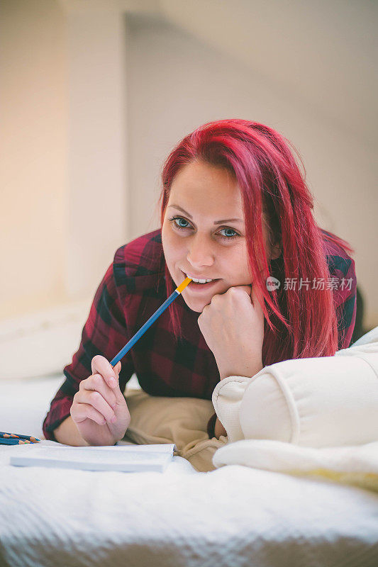 微笑的漂亮的年轻女孩躺在一个舒适的床上，她的纸，颜料和蜡笔为美术课