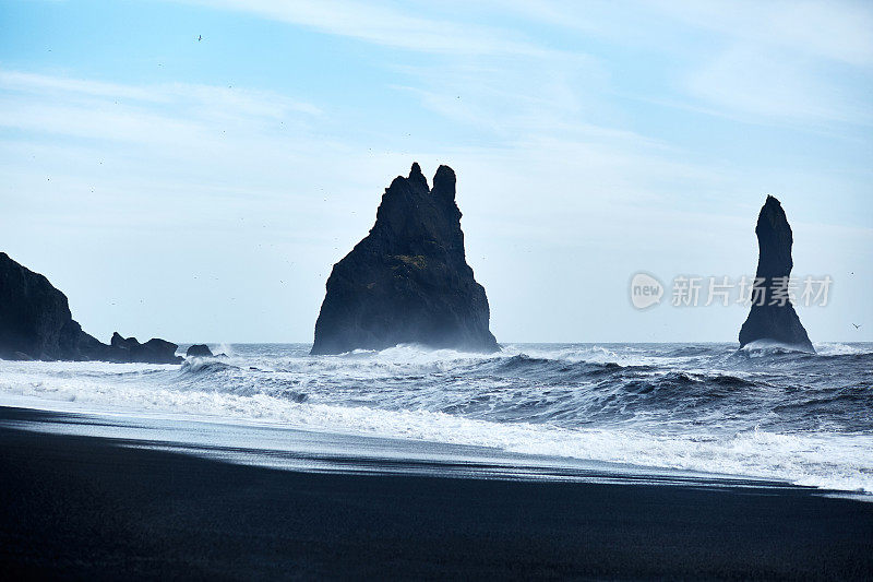 风景优美的黑色岩层突出沉重的海洋。在蓝天的背景下，强大的海浪撞击着黑色的火山沙滩。湾、冰岛