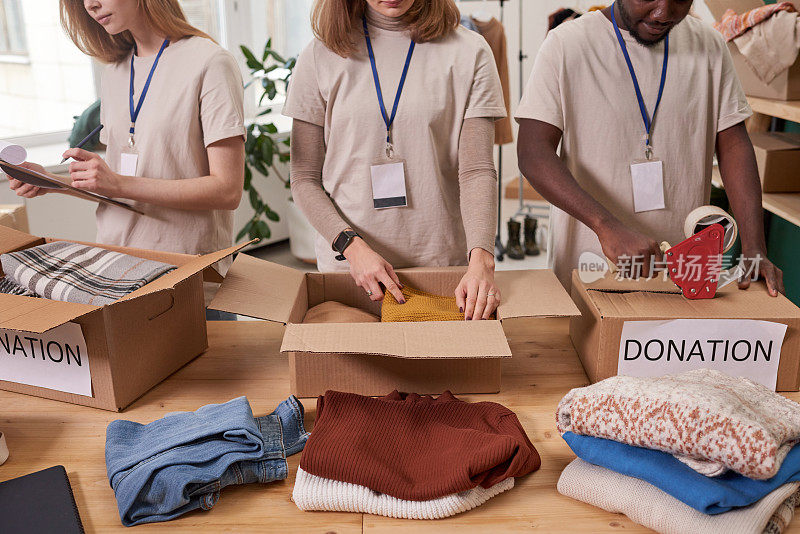 高角度拍摄三名身穿米色t恤将衣物装进纸板箱进行捐赠的志愿者
