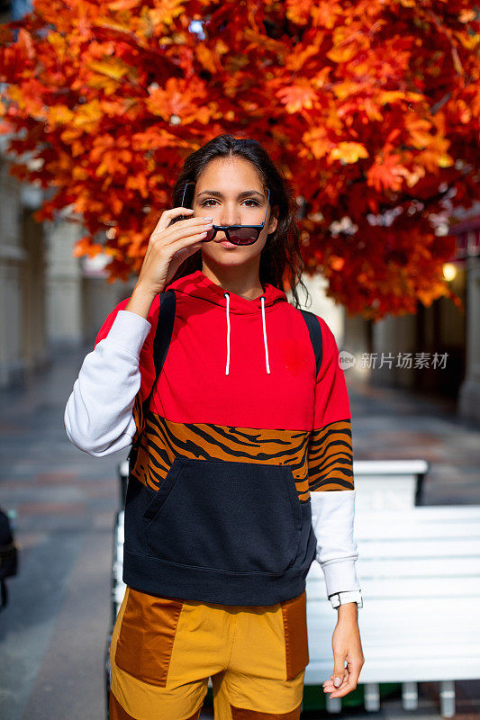 美丽的年轻女子在红色的运动服脱下她的太阳镜，站在树与茂盛的红色树叶