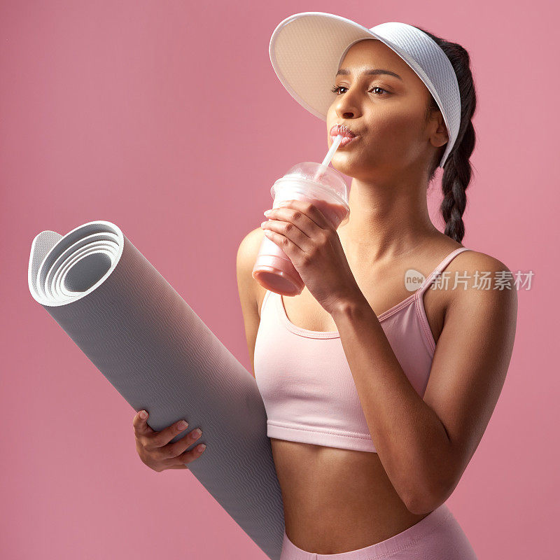 一个迷人和运动的年轻女子摆姿势与一个运动垫和冰沙在工作室以粉红色的背景
