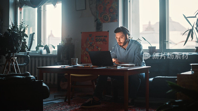 年轻英俊的男人带着耳机在创意公司的笔记本电脑上工作。装修时尚的装饰与室内植物，艺术海报和大圆形窗户。