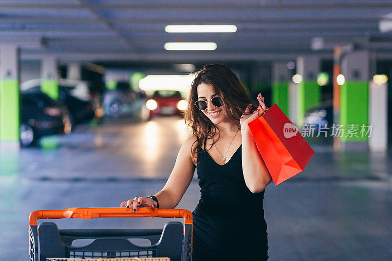 年轻漂亮的女子推着购物车，拿着装着商品和物品的包裹，来到购物中心地下停车场的汽车前