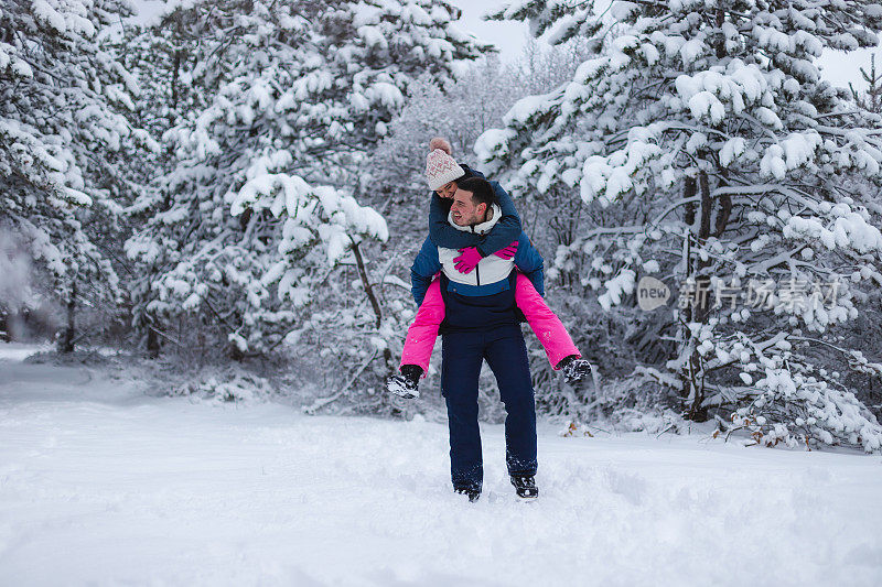 年轻的男朋友背着他的女朋友在雪地里玩，周围是雪树