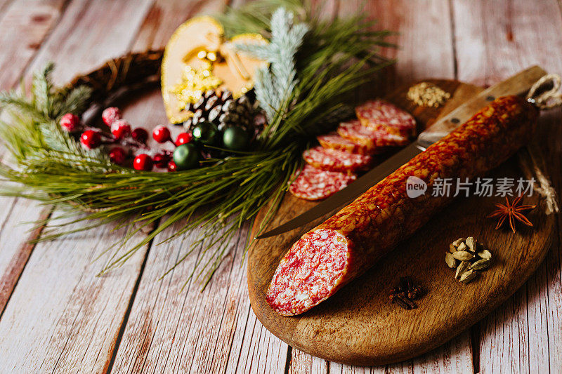 一份自制的意大利腊肠放在木制砧板上，配以豆蔻、丁香、八角、菜刀和一个闪闪发光的木制心形圣诞装饰