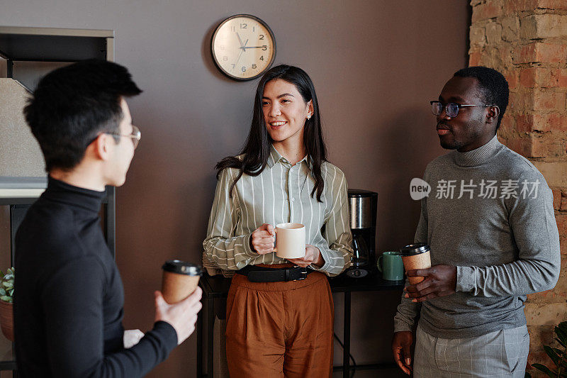 水平拍摄三个多民族的人站在阁楼办公室里，边喝咖啡边聊着什么