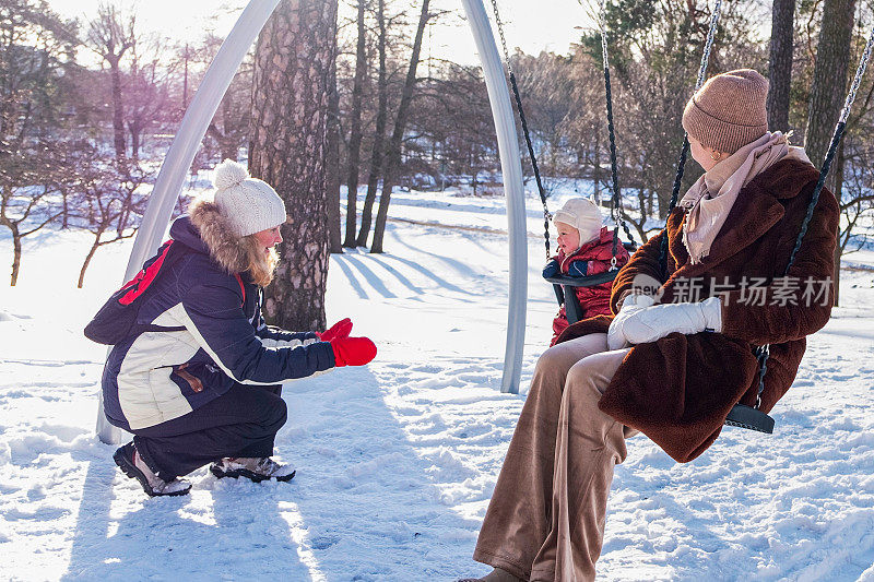年轻的白人妇女带着孩子在操场上的冬季公园散步。和朋友一起家庭度假。