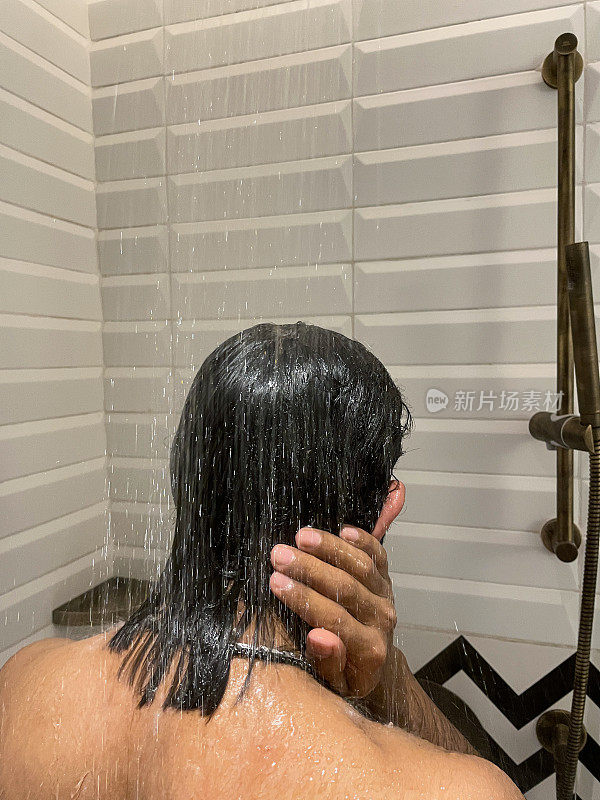 印度男子在淋浴间洗头，在流水下冲洗头发上的肥皂泡沫的特写，白色斜边墙砖呈水平堆叠图案，后视图，焦点在前景