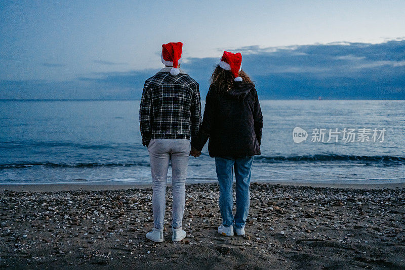 希腊，一对戴着圣诞帽的年轻夫妇在黎明时分欣赏着海岸线的景色