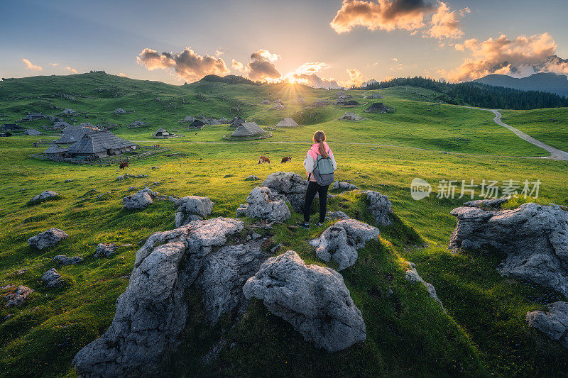 夏日夕阳下，美丽古老的高山村庄里，石头上背着背包的女孩，绿色草地上的奶牛。有年轻女人的风景，房子，树木，天空。旅行和徒步旅行。斯洛文尼亚