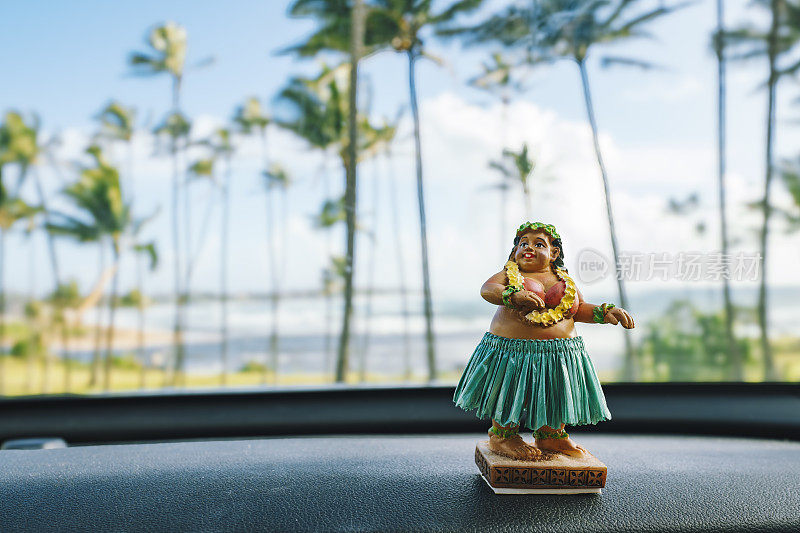 夏威夷草裙舞舞者娃娃坐在汽车仪表板上，背景是海滩和棕榈树