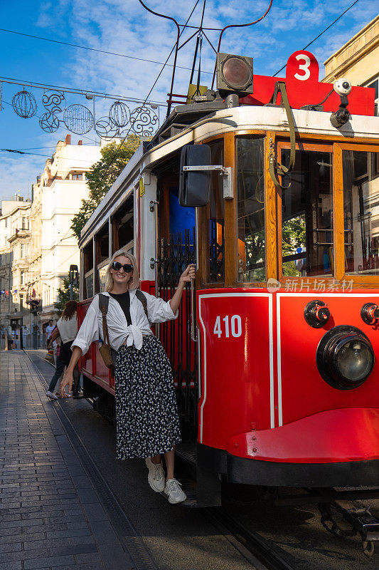 土耳其伊斯坦布尔，Istiklal大街上，一辆怀旧的红色Istiklal电车的运行板上挂着一个背包，穿着白衬衫和裙子的年轻金发美女。垂直图
